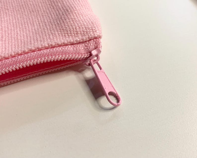 Puede incluir: Primer plano de una bolsa rosa con cremallera. La bolsa est&aacute; hecha de una tela texturizada y presenta una cremallera rosa a juego. El tirador de la cremallera tambi&eacute;n es rosa y tiene forma redondeada.