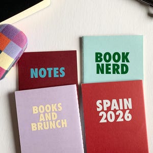 May include: Four small rectangular notebooks with coloured covers and text. One is burgundy with blue 'NOTES' text, one is light blue with green 'BOOK NERD' text, one is lavender with cream 'BOOKS AND BRUNCH' text, and one is red with white 'SPAIN 2026' text.
