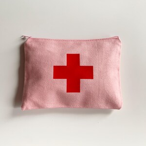 Puede incluir: Un estuche rosa de lona con cremallera y una gran cruz roja en el centro. El estuche rectangular parece estar hecho de una tela texturizada. La cruz roja es de un color s&oacute;lido y llamativo, que contrasta con el fondo rosa.