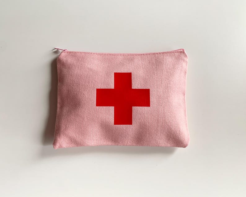Puede incluir: Un estuche rosa de lona con cremallera y una gran cruz roja en el centro. El estuche rectangular parece estar hecho de una tela texturizada. La cruz roja es de un color s&oacute;lido y llamativo, que contrasta con el fondo rosa.