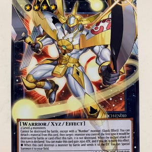 Puede incluir: Una carta coleccionable de "Número 39 Utopia el Enviado de la Luz". Muestra un guerrero con armadura blanca y dorada, empuñando dos espadas. El texto describe sus habilidades y estadísticas, sobre un fondo azul oscuro.