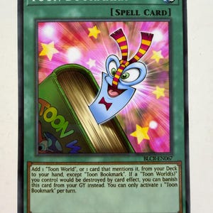 May include: A Yu-Gi-Oh! trading card titled Toon Bookmark, a green Spell Card featuring a blue cartoon character emerging from a green book against a pink background with yellow stars. The card text details game mechanics and card identification.