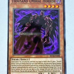 Peut inclure: Une carte à collectionner Yu-Gi-Oh! intitulée Thousand Umbral Horror, illustrant une créature ailée en armure violet foncé. La carte affiche le texte Fiend/Effect, ATK 0, DEF 0, le code BETB-EN008 et quatre étoiles jaunes dans la partie supérieure.