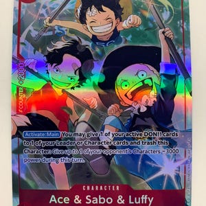 May include: A holographic trading card featuring three animated characters: Ace, Sabo, and Luffy. The card has a red border with the number 1 and text that reads "1000 Strike". The card also includes the text "COUNTER +2000" and "Goa Kingdom".