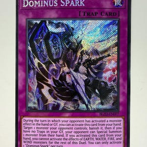 Op de afbeelding: Een holografische Dominus Spark-kaart met een paarse rand. De kaart toont een gedetailleerde illustratie van een personage met donkere bepantsering en een dynamische pose. Tekst bevat "DOMINUS SPARK" en "TRAP CARD", samen met spelinstructies.