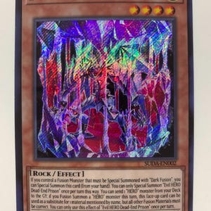 May include: A holographic trading card featuring the text "EVIL HERO DEAD-END PRISON." The card has a dark, multi-colored background with a central image of a monster. The card's border is brown, and it includes text detailing its effects and stats.