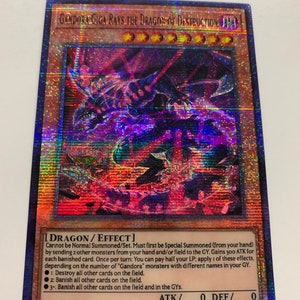 Peut inclure: Une carte de jeu de commerce holographique repr&eacute;sentant un dragon noir et rouge avec des yeux rouges brillants. La carte est intitul&eacute;e "Gandora Giga Rays the Dragon of Destruction" et a un fond noir avec une bordure bleue et violette. La carte est une carte "Dragon/Effet" avec une puissance d'attaque de 0 et une puissance de d&eacute;fense de 0. Le texte de la carte se lit comme suit : "Ne peut pas &ecirc;tre Invoqu&eacute; Normalement/Plac&eacute;. Doit d'abord &ecirc;tre Invoqu&eacute; Sp&eacute;cialement (de votre main) en envoyant 2 autres monstres de votre main et/ou du terrain au Cimeti&egrave;re. Gagne 300 ATK pour chaque carte bannie. Une fois par tour : Vous pouvez payer la moiti&eacute; de vos PV ; appliquez l'un de ces effets, en fonction du nombre de monstres "Gandora" avec des noms diff&eacute;rents dans votre Cimeti&egrave;re. 1 : D&eacute;truisez toutes les autres cartes sur le terrain. 2 : Bannissez toutes les autres cartes sur le terrain. 3 : Bannissez toutes les autres cartes sur le terrain et dans les Cimeti&egrave;res."