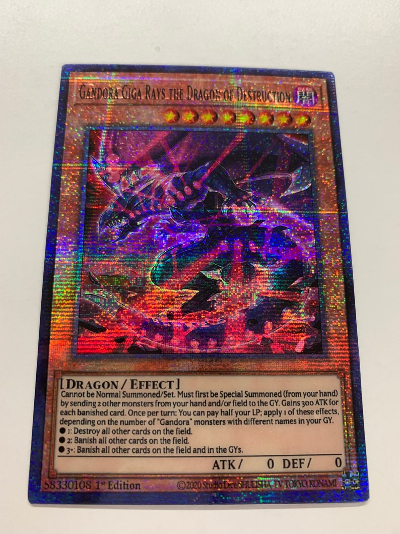 Peut inclure: Une carte de jeu de commerce holographique repr&eacute;sentant un dragon noir et rouge avec des yeux rouges brillants. La carte est intitul&eacute;e "Gandora Giga Rays the Dragon of Destruction" et a un fond noir avec une bordure bleue et violette. La carte est une carte "Dragon/Effet" avec une puissance d'attaque de 0 et une puissance de d&eacute;fense de 0. Le texte de la carte se lit comme suit : "Ne peut pas &ecirc;tre Invoqu&eacute; Normalement/Plac&eacute;. Doit d'abord &ecirc;tre Invoqu&eacute; Sp&eacute;cialement (de votre main) en envoyant 2 autres monstres de votre main et/ou du terrain au Cimeti&egrave;re. Gagne 300 ATK pour chaque carte bannie. Une fois par tour : Vous pouvez payer la moiti&eacute; de vos PV ; appliquez l'un de ces effets, en fonction du nombre de monstres "Gandora" avec des noms diff&eacute;rents dans votre Cimeti&egrave;re. 1 : D&eacute;truisez toutes les autres cartes sur le terrain. 2 : Bannissez toutes les autres cartes sur le terrain. 3 : Bannissez toutes les autres cartes sur le terrain et dans les Cimeti&egrave;res."