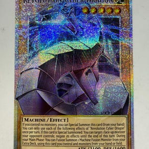Könnte beinhalten: Holografische Sammelkarte mit "Revolution Cyber Dragon". Die Karte hat ein blaues und silbernes Farbschema mit einem futuristischen Drachendesign. Der Kartentext enthält "MACHINE / EFFECT", ATK/2100 und DEF/1600. Eine Sammelkarte für Fans.