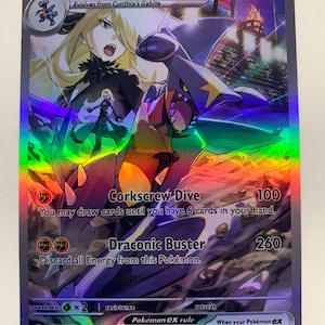 Puede incluir: Carta coleccionable Pokémon holográfica con Cynthia's Garchomp EX. La tarjeta muestra una colorida ilustración de una mujer rubia y un Garchomp. El texto incluye "Corkscrew Dive" y "Draconic Buster" con valores de daño.