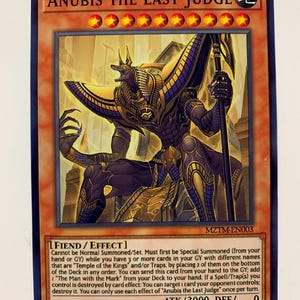 May include: Anubis the Last Judge trading card from the Yu-Gi-Oh! Trading Card Game. The card is a "FIEND/EFFECT" monster with an attack power of 3000 and a defense power of 0. The card features an illustration of Anubis, the Egyptian god of the dead, in a golden and purple outfit, holding a staff. The card is set against a background of a golden temple.