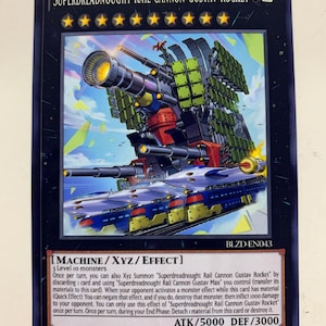 May include: A trading card featuring the Superdreadnought Rail Cannon Gustav Rocket. The card depicts a large, detailed war machine with multiple cannons, in shades of blue, green, red, and yellow. The card has text that includes ATK/5000 DEF/3000.