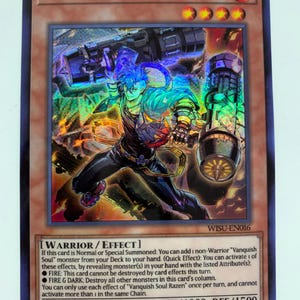 May include: A holographic trading card featuring "Vanquish Soul Razen." The card shows a character with blue hair and armour, wielding futuristic weaponry. The card's text includes game effects, attack, and defence stats. The card is a 1st Edition.
