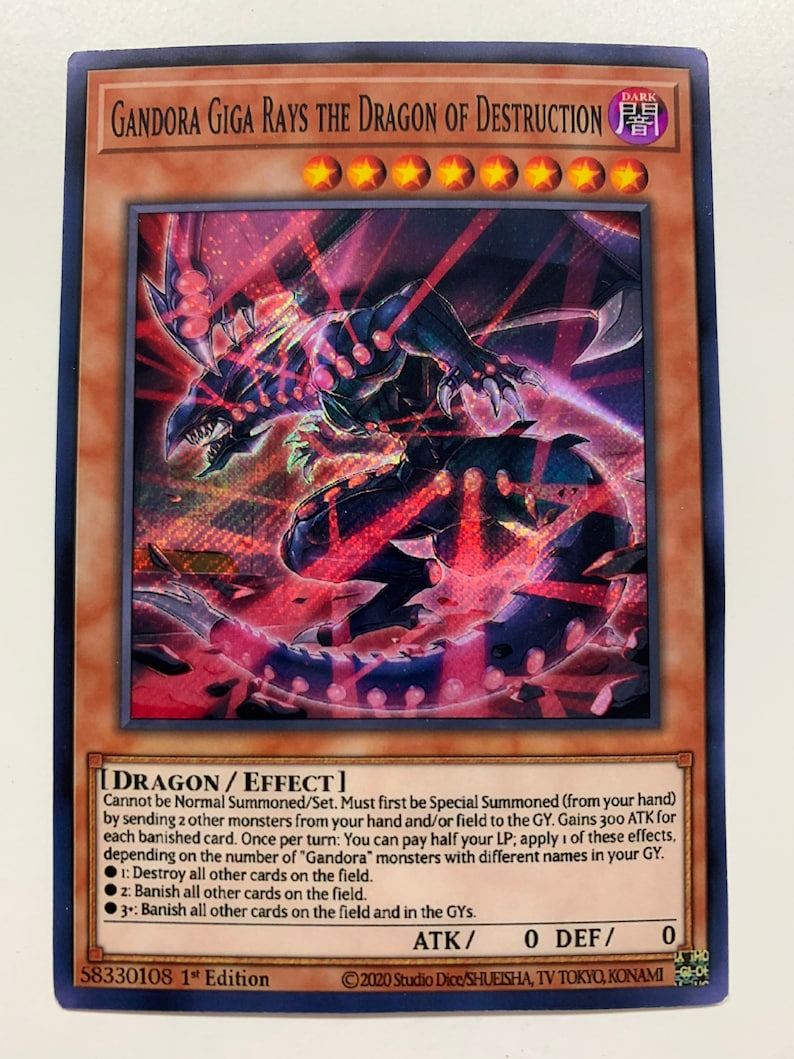 Peut inclure: Une carte de jeu de cartes &agrave; collectionner Yu-Gi-Oh! pour le monstre "Gandora Giga Rays the Dragon of Destruction". La carte repr&eacute;sente un dragon sombre aux yeux rouges et au corps noir et rouge. La carte a un fond sombre avec une bordure rouge et noire. Le texte de la carte se lit comme suit : "Ne peut pas &ecirc;tre Invoqu&eacute; Normalement/Plac&eacute;. Doit d'abord &ecirc;tre Invoqu&eacute; Sp&eacute;cialement (depuis votre main) en envoyant 2 autres monstres de votre main et/ou du terrain au Cimeti&egrave;re. Gagne 300 ATK pour chaque carte bannie. Une fois par tour : Vous pouvez payer la moiti&eacute; de vos PV ; appliquez 1 de ces effets, en fonction du nombre de monstres "Gandora" avec des noms diff&eacute;rents dans votre Cimeti&egrave;re. 1 : D&eacute;truisez toutes les autres cartes sur le terrain. 2 : Bannissez toutes les autres cartes sur le terrain. 3+ : Bannissez toutes les autres cartes sur le terrain et dans les Cimeti&egrave;res."