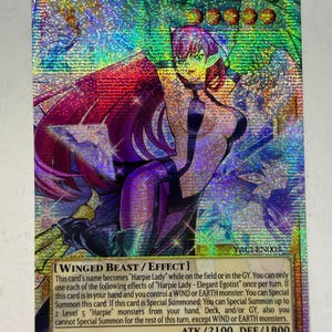 Puede incluir: Una carta coleccionable holográfica de Yu-Gi-Oh! titulada Harpie Lady - Elegant Egotist. La carta muestra un personaje de pelo morado con grandes alas, vistiendo un traje oscuro. El texto incluye Winged Beast/Effect, ATK/2100 y DEF/1800.