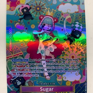 May include: A holographic trading card featuring the character Sugar from Donquixote Pirates. The card has a colorful design with a central illustration of Sugar, a crown, and bear-like figures. Text includes "Sugar" and "Donquixote Pirates".