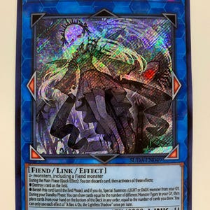 Puede incluir: Una carta de juego de colección « A Bao A Qu, The Lightless Shadow » azul, plateada y negra que representa a un personaje femenino con el pelo largo blanco y un vestido oscuro. La carta es una carta « Fiend/Link/Effect » con una potencia de ataque de 2800 y un rango de enlace de 4. El texto de la carta describe las capacidades y los efectos de la carta.