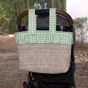 May include: A rectangular stroller organizer bag featuring a textured tan and brown woven body with a light green and white gingham patterned fabric trim, a ruffled top edge, and two matching gingham fabric straps for attachment to a handle.