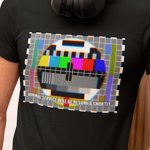 May include: Black t-shirt with a retro test card graphic, including the text "NORMAL SERVICE WILL BE RESUMED SHORTLY." The graphic displays a range of colours and shapes. The t-shirt has a crew neck design.