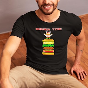 Könnte beinhalten: Schwarzes T-Shirt mit einem pixeligen Burger-Motiv und dem roten Text "BURGER TIME". Der Burger hat Schichten aus Brötchen, Salat, Tomate und einem Patty, mit einem pixeligen Koch oben drauf. Das Shirt hat einen Rundhalsausschnitt.