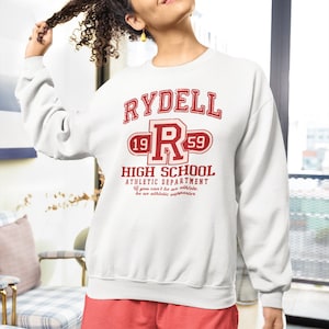 Puede incluir: Sudadera blanca con el texto "RYDELL HIGH SCHOOL ATHLETIC DEPARTMENT" en rojo. La sudadera también presenta una gran "R" roja con los números "19" y "59". La persona lleva pantalones cortos rojos.