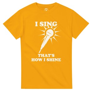 May include: Mustard yellow t-shirt with a white graphic of a microphone radiating sunbeams. The text "I SING" is above the microphone, and "THAT'S HOW I SHINE" is below. The shirt is made of a soft material.