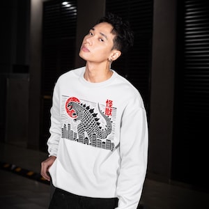 Può includere: Felpa bianca con grafica nera e rossa di una creatura simile a Godzilla, un sole nascente e un paesaggio urbano. Il testo giapponese è anch'esso nero. Maglietta a maniche lunghe con girocollo.