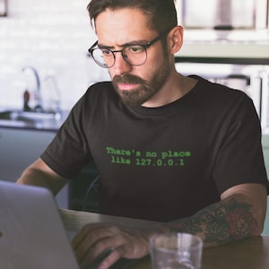 Puede incluir: Un hombre con una camiseta negra con el texto "There's no place like 127.0.0.1" en texto verde. Está sentado en una mesa con un portátil y un vaso de líquido.