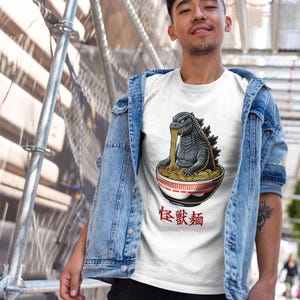 May include: A white t-shirt with a graphic of a Godzilla-like monster eating ramen noodles from a bowl. The text "怪獣麺" is printed below the bowl.