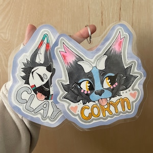 May include: Two cartoon-style character badges. One features a black and white wolf with the name "CLAY" in silver lettering. The other has a black, blue, and pink wolf with the name "CORYN" in gold lettering. Both have a light blue border.