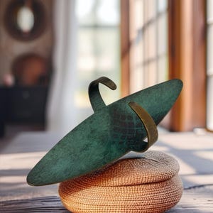 May include: A teal-colored, elongated sculpture with bronze accents, resting on a woven basket. The sculpture has a textured surface and a curved bronze band. The basket is a light brown color and has a round shape. The sculpture is on a wooden surface.