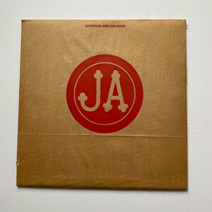 May include: A brown cardboard record album cover with the text "JEFFERSON AIRPLANE/BARK" printed in white at the top. A red circle with the letters "JA" in white is in the center of the cover.