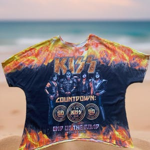 Op de afbeelding: Zwart T-shirt met de band KISS en een vurig ontwerp. Het shirt toont de naam van de band in goud, de woorden "COUNTDOWN: END OF THE ROAD WORLD TOUR" en de bandleden. Het shirt heeft een tie-dye effect met oranje en gele vlammen.