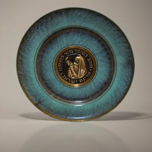 May include: A decorative plate with a turquoise and bronze finish. The plate features a central medallion with a religious figure and Hebrew text. The plate has a circular design with concentric rings.