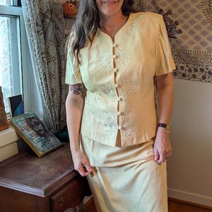 May include: A pale yellow two-piece skirt suit featuring a short-sleeved button-down jacket with white floral embroidery along the front placket and a matching knee-length skirt. The fabric has a subtle tonal pattern throughout the garment.