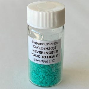 May include: A small glass vial with a white cap, filled with bright turquoise granules. The label reads "Copper Chloride CuCl2-(H2O)2 NEVER INGEST! TOXIC TO HEALTH!" and "SilverGal LLC".