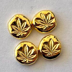 May include: Four gold-colored, irregularly shaped buttons with a cannabis leaf design. The buttons have a shiny, reflective surface and are arranged on a light gray background. The leaf design is etched into the surface.