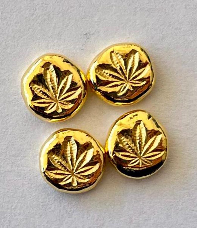 May include: Four gold-colored, irregularly shaped buttons with a cannabis leaf design. The buttons have a shiny, reflective surface and are arranged on a light gray background. The leaf design is etched into the surface.