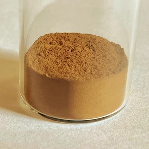 May include: A clear glass vial containing a finely ground, medium-brown powder. The powder has a matte, textured surface and fills the bottom portion of the cylindrical container, showing a consistent earthy tone throughout the substance.