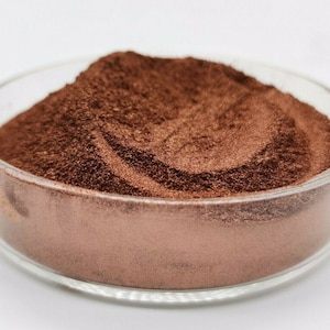 Puede incluir: Primer plano de un pequeño plato lleno de una gran cantidad de polvo fino de color marrón oscuro. El polvo tiene un ligero brillo metálico y parece ser un pigmento o un colorante.