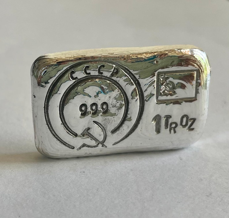 May include: A silver bullion bar with the inscription "1 Tr Oz" and "999" in a circle with a hammer and sickle symbol. The bar also has the inscription "CCCP" above the circle.