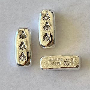 May include: Three silver bars with the Freemason symbol. Two bars are rectangular with rounded edges, each displaying three symbols. The third bar is smaller, rectangular, and has the text "10 GRAM 999FS" and a single symbol.