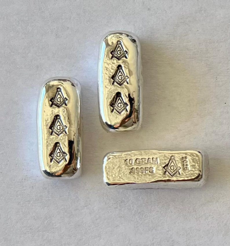 May include: Three silver bars with the Freemason symbol. Two bars are rectangular with rounded edges, each displaying three symbols. The third bar is smaller, rectangular, and has the text "10 GRAM 999FS" and a single symbol.