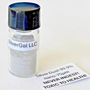 Puede incluir: Un pequeño frasco de vidrio con tapa negra que contiene un polvo gris. La etiqueta en el frasco dice "SilverGal LLC". Una etiqueta blanca en la mesa frente al frasco dice "Silver Dust 99.9% nano-75µm ¡NUNCA INGERIR! ¡TÓXICO PARA LA SALUD!"