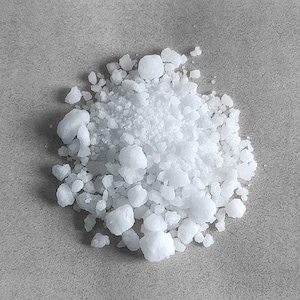 May include: A close-up shot of a pile of white, crystalline substance. The crystals vary in size, with some appearing as small granules and others as larger, irregular chunks. The background is a plain, light gray surface.