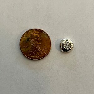 K&ouml;nnte beinhalten: Ein runder Metall-Token in Silberfarbe mit einem eingegravierten Freimaurer-Symbol. Der Token ist etwa so gro&szlig; wie ein Cent-St&uuml;ck.