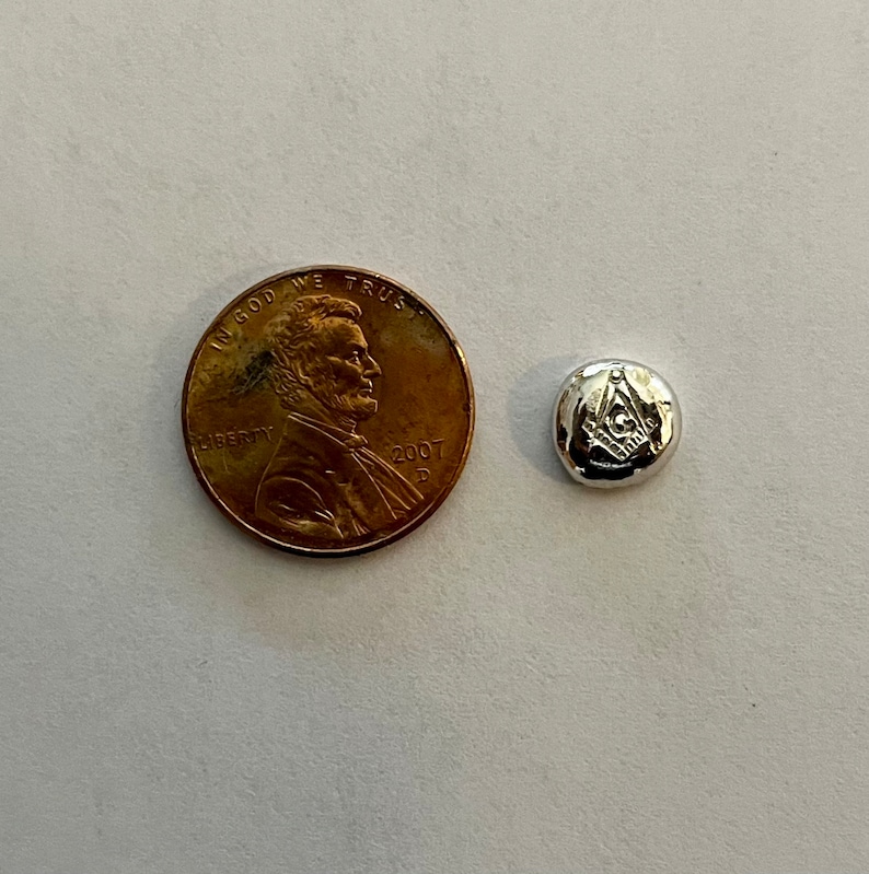 K&ouml;nnte beinhalten: Ein runder Metall-Token in Silberfarbe mit einem eingegravierten Freimaurer-Symbol. Der Token ist etwa so gro&szlig; wie ein Cent-St&uuml;ck.