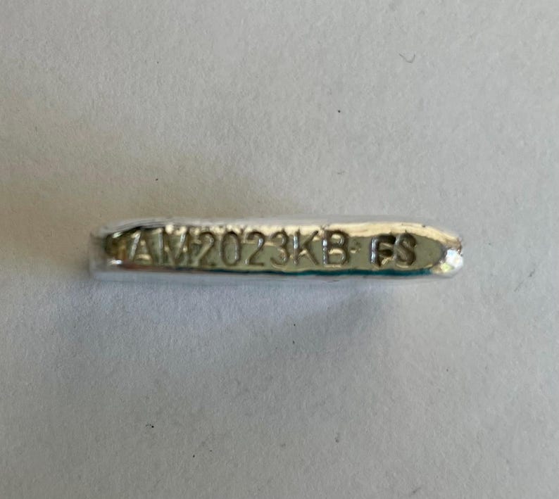 May include: A silver bar with the inscription "AM2023KB FS" on it.