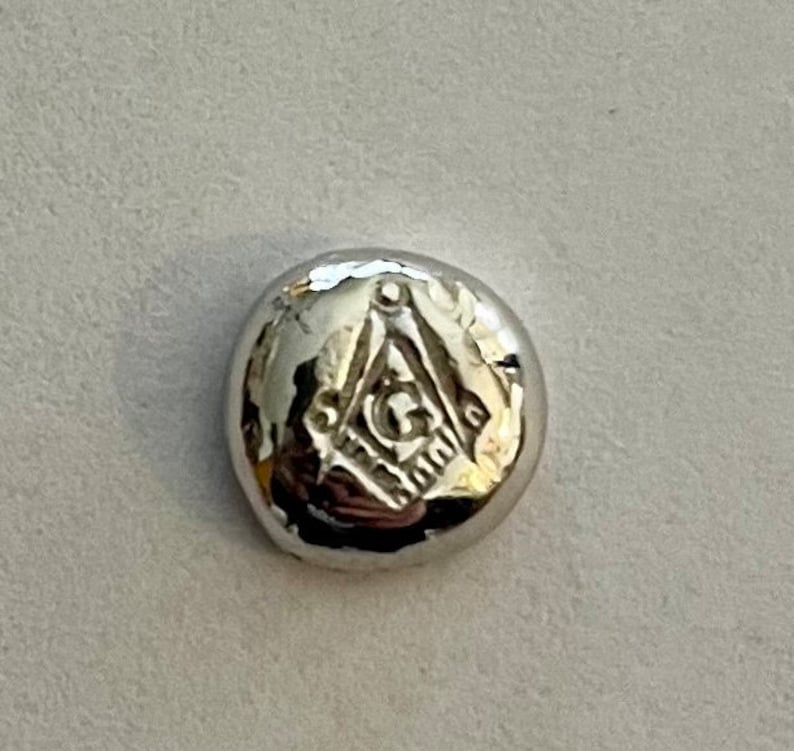 K&ouml;nnte beinhalten: Ein runder Anh&auml;nger aus silberfarbenem Metall mit einem erhabenen freimaurerischen Symbol, bestehend aus einem Zirkel und einem Winkelma&szlig; mit einem 'G' in der Mitte.