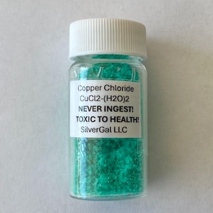 Può includere: Una piccola bottiglia di vetro con tappo bianco piena di cristalli verde brillante. Un'etichetta bianca sulla bottiglia recita "Cloruro di rame" e avverte "TOSSICO PER LA SALUTE!" Il prodotto è di SilverGal LLC.